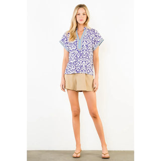 A woman modeling a short-sleeve button-front top with a blue and white geometric pattern, paired with tan shorts and sandals, standing against a plain white studio background.