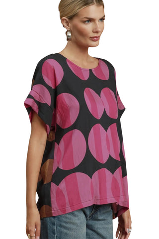 A woman wearing a short-sleeve black top with a bold pink and magenta abstract circular pattern, paired with blue jeans, against a plain white background.
