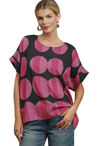 A woman modeling a short-sleeve top with a bold black and pink circular dot pattern, paired with blue jeans, against a plain white background.