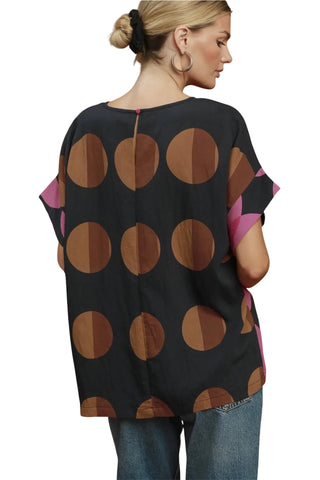 A woman shown from the back wearing a loose black top with large brown polka dots and pink side panels, paired with blue jeans, against a plain white background.