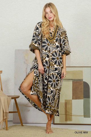 A woman modeling a loose, knee-length kaftan dress with a bold floral and paisley print in olive green, black, and cream. The dress features a V-neckline with a gold-toned trim and wide, elbow-length sleeves.