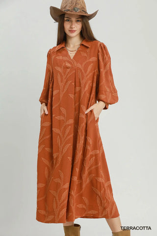 A woman wearing a terracotta-colored midi shirt dress with a tonal leaf pattern, paired with a brown cowboy hat and boots, against a light gray studio background.