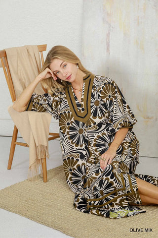 A woman models a loose, V-neck kaftan dress with a bold floral pattern in olive green, black, and cream. She is seated on a woven rug next to a wooden chair with a draped beige throw blanket, against a textured white wall.