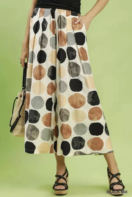 A model wearing wide-leg culottes with a large watercolor-style polka dot pattern in black, brown, beige, and gray on an off-white base, paired with black wedge sandals and holding a woven handbag against a solid light green background.
